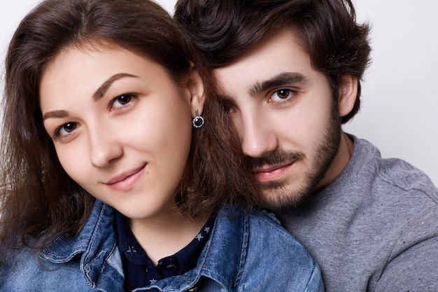 A close-up of two young people in love over white wall. A bearded hipster embracing her girlfriend with charming eyes and smile