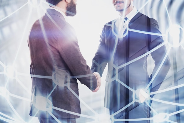 Close up of two young unrecognizable businessmen shaking hands over skyscraper background with double exposure of people network hologram. Concept of partnership. Toned image