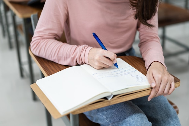 Primo piano degli studenti universitari che scrivono una nota sul taccuino in classe mentre ascoltano e studiano la lezione. primo piano delle mani della donna che scrivono su un blocco note posizionato su una scrivania in legno