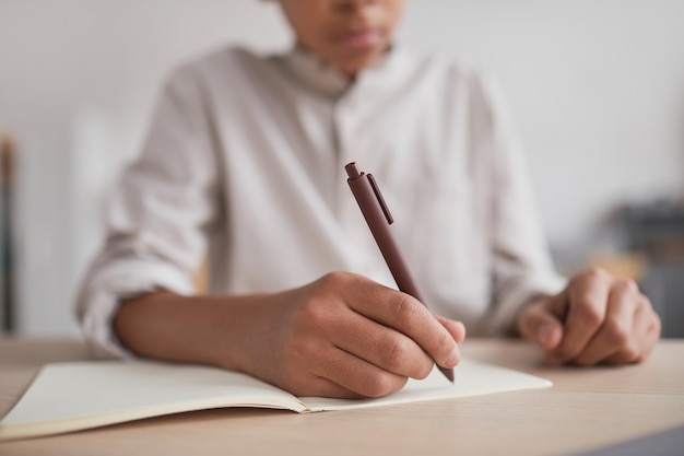 Close up of unrecognizable African-American boy doing homework and writing in notebook while sitting at desk, focus on hand holding pen, copy space