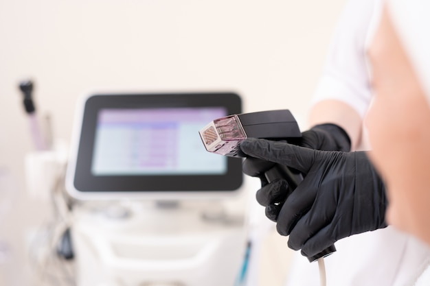 Close-up of unrecognizable beautician in black latex gloves pointing at handle of facial machine while explaining client how it works