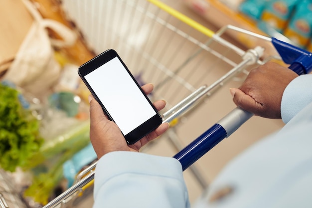 Primo piano di una donna nera irriconoscibile che acquista generi alimentari nel supermercato e utilizza lo smartphone con whi