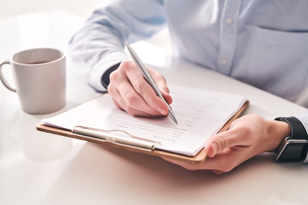 Close-up of unrecognizable businessman sitting at table with tea mug and making notes in application form
