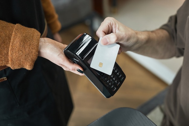 Photo close-up of unrecognizable customer paying with contactless card and putting it to terminal held by waitress in cafe