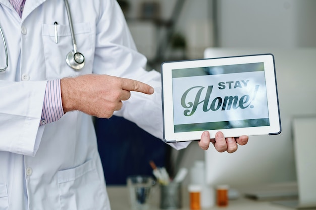 Close-up of unrecognizable doctor in white coat pointing at tablet with Stay home text