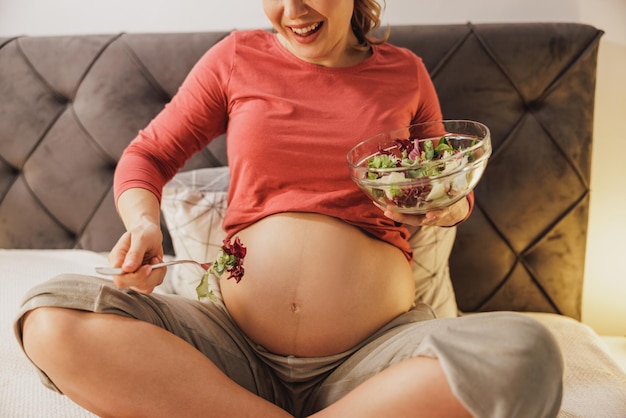 Close up of an unrecognizable happy expecting woman eating fresh vegetable salad while sitting on the bed.