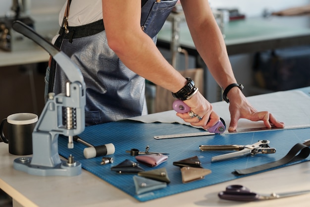 Close-up of unrecognizable man in apron using metal ruler to cut edge of leather in craft studio