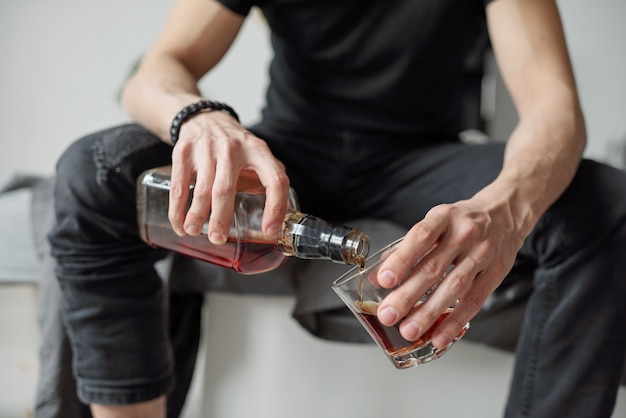 Close-up of unrecognizable man in black jeans pouring whisky into glass while drinking alone