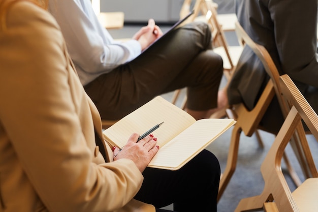 Photo close up of unrecognizable mature businesswoman holding open note pad while sitting in audience during conference or seminar, copy space