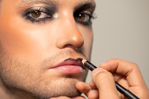 Close-up of unrecognizable MUA professional making lip contour with black liner to young bearded man against isolated background