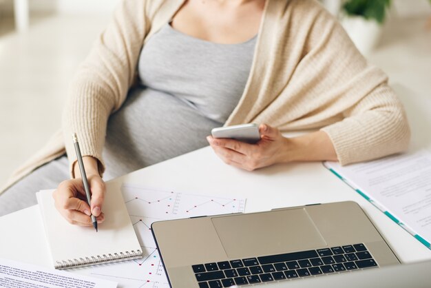 Photo close-up of unrecognizable pregnant businesswoman with smartphone in hand sitting at table and making notes about tasks while viewing data on laptop