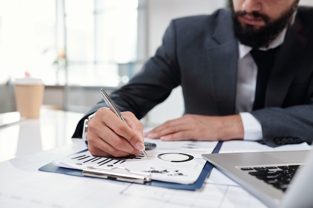 Close up of unrecognizable successful businessman writing on clipboard filling data graphs and charts while working in office , copy space