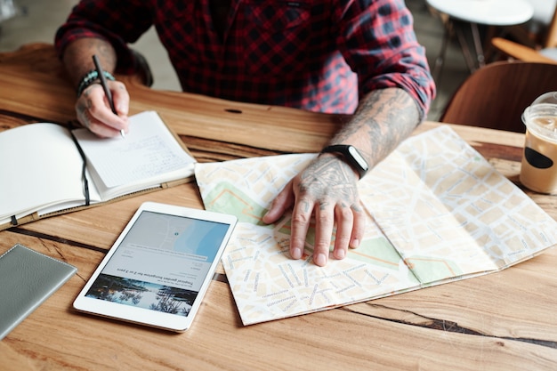 Photo close-up of unrecognizable tattooed man sitting at table in cafe and checking hotel location on map while planning trip