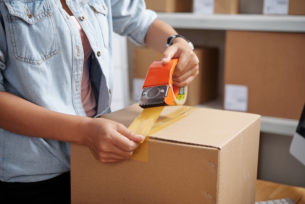 Close-up of unrecognizable woman in denim shirt using duct tape\
while packaging cardboard box