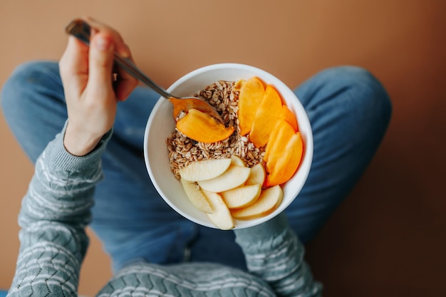 Close up unrecognizable woman hand holds a spoon with a piece of bitten fruit over a plate of muesli