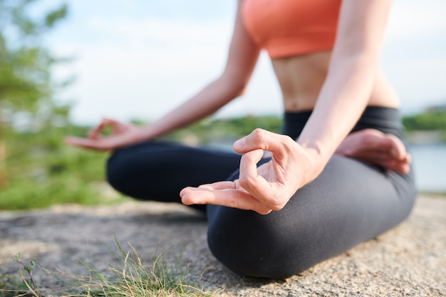 Primo piano di una donna irriconoscibile in leggings che si siede nella posizione del loto e tiene le mani nel mudra durante la meditazione all'aperto