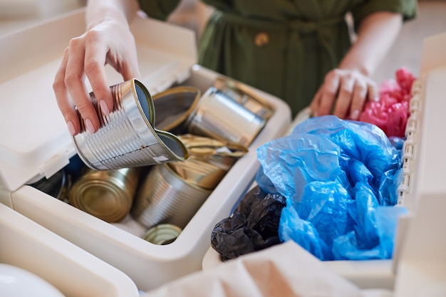 Close up of unrecognizable woman putting discarded metal cans into trash bin while sorting waste at home