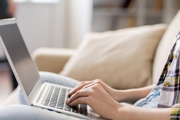 Close-up of unrecognizable woman sitting on comfortable sofa and typing on laptop