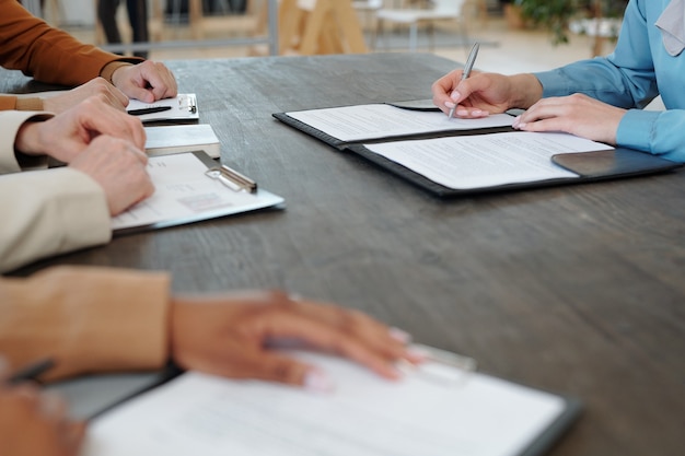 Close-up of unrecognizable woman sitting at table in front of company representatives and signing contract with company after interview