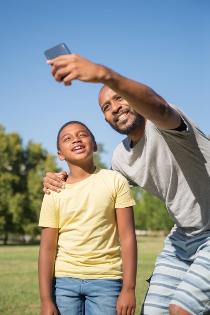 Close-up van Afro-Amerikaanse jongen en zijn vader met telefoon. Glimlachende bebaarde man met telefoon en zijn zoon die dichtbij staan, beide kijkend naar camera die selfie neemt. Vrije tijd en modern technologieconcept