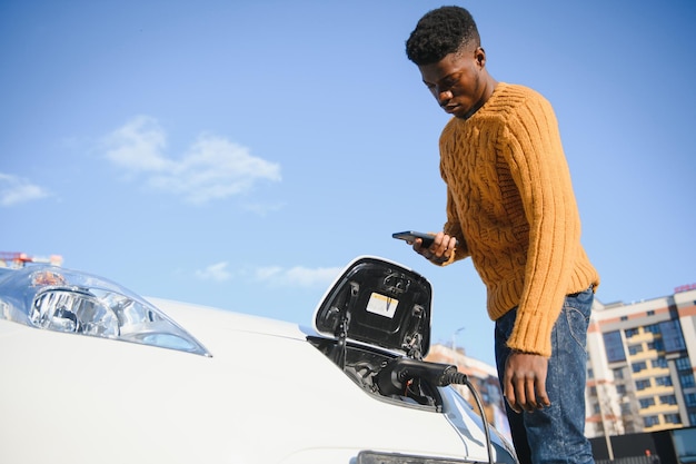 Close up van Afro-Amerikaanse man oplaadkabel aansluiten op elektrische auto. Jonge man permanent in de buurt van zijn moderne auto met lederen koffer in de hand.