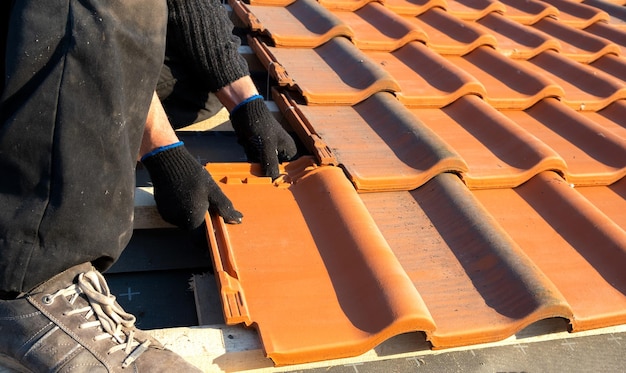 Close-up van arbeidershanden die gele keramische dakpannen installeren die op houten planken zijn gemonteerd die het dak van een woongebouw in aanbouw bedekken.