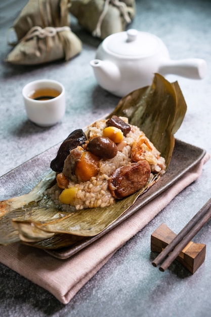 Close up van Aziatische smakelijke zelfgemaakte gerechten in drakenboot (duan wu) festival, rijst knoedels of zongzi verpakt door gedroogde bamboe bladeren op plaat met thee op zwarte ondergrond