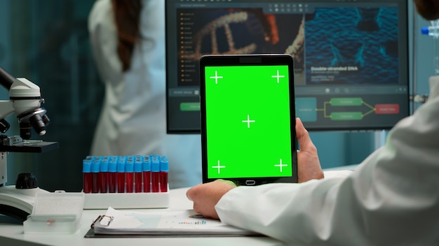 Close up van biochemicus zittend op de werkplek in laboratorium met behulp van groene mock-up scherm tablet met chroma key display. collega die op de achtergrond werkt en bloedmonsters brengt.