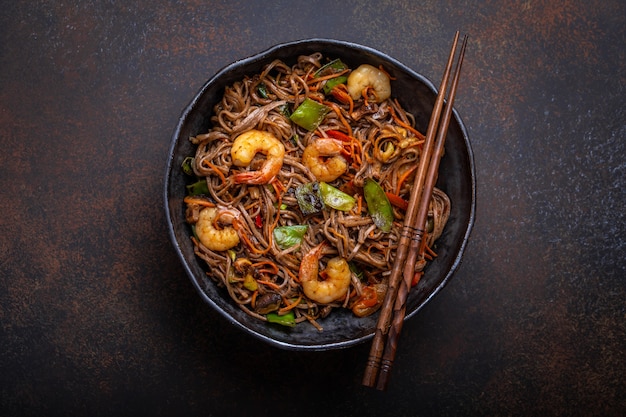 Close-up van Chinese soba roerbak noedels met garnalen, groenten in rustieke keramische kom pan geserveerd op betonnen achtergrond, close-up, bovenaanzicht. Traditioneel Aziatisch/Thaise gerecht, Chinese maaltijd