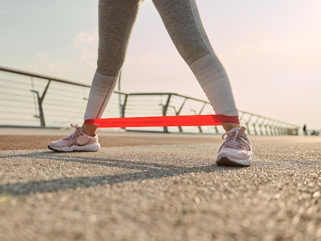 Close-up van de benen van een atleetvrouw, sportvrouw in grijze leggins die lichaamsgewichttraining doen, trainen met een elastische weerstandsband op een stadsbrug bij zonsopgang