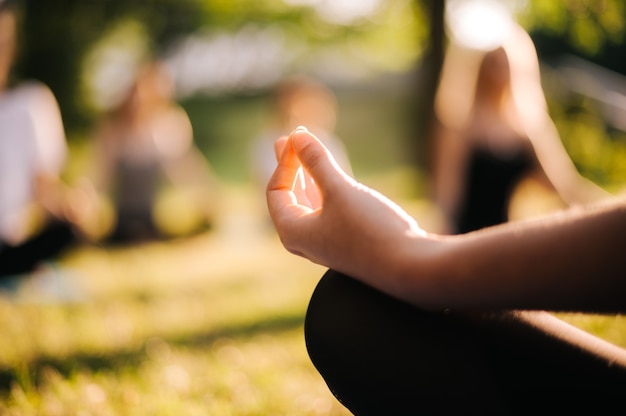 Close-up van de hand van een jonge yogi-vrouw die in de lotushouding zit in het stadspark op zonnige zomerochtend bij zonsopgang. meisje mediteert met vingers in jnana mudra. wazige achtergrond van groep mediterende mensen