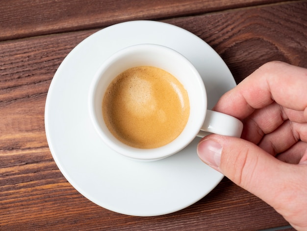 Close-up van de hand van een man die een klein wit kopje aromatische espresso vasthoudt op een witte schotel. Houten achtergrond, bovenaanzicht, plat leggen