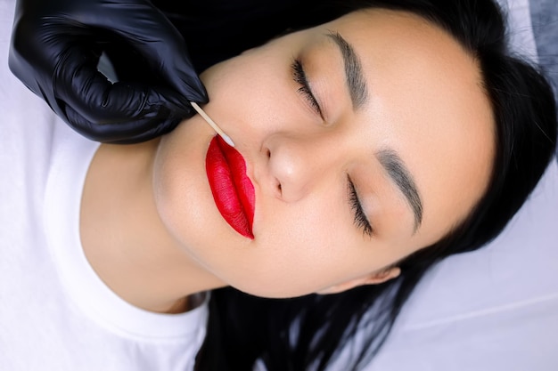 Close-up van de hand van een permanente make-upmeester die overtollige rode lippenstift van de lippen verwijdert met een wattenstaafje vóór de tatoegeringprocedure