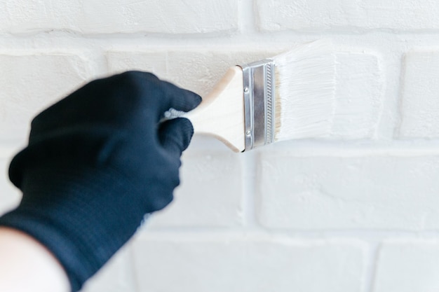 Close-up van de hand van een werknemer met een borstel die een bakstenen muur schildert met witte verf