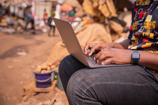 Close-up van de handen van een Afrikaanse jongen die een laptop gebruikt terwijl hij op de markttechnologie in Afrika zit