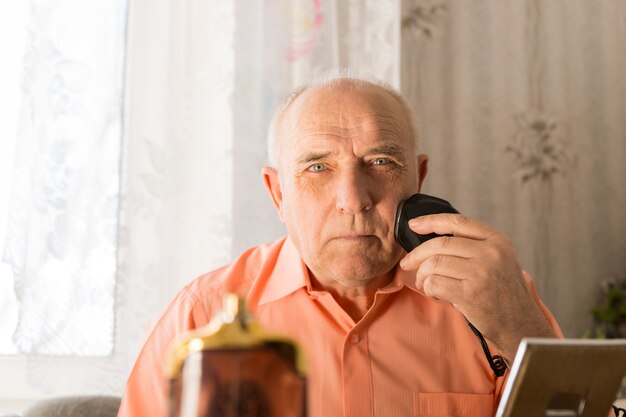 Close-up van de oude man die zijn haar op het gezicht scheert met een elektrisch scheerapparaat terwijl hij naar de camera kijkt.