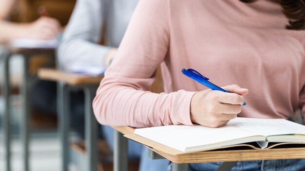 Close-up van de universiteitsstudenten die een notitie schrijven op een notitieboekje in de klas terwijl ze luisteren en de lezing bestuderen. Close up van de handen van de vrouw schrijven op Kladblok geplaatst op houten bureaublad