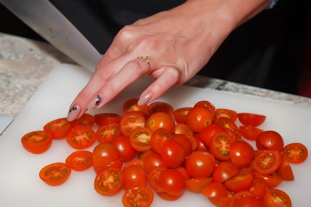 Close-up van de vrouw van de chef-kok die cherrytomaatjes snijdt met een mes op plastic witte snijplank in de thuiskeuken. Achtergrond voor het koken. Concept van culinaire uitmuntendheid en catering. Ruimte kopiëren