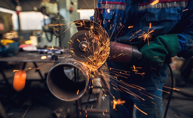 Close-up van de zijkant van professionele hardwerkende man in uniform snijdt metalen pijp sculptuur met een grote elektrische slijper terwijl vonken vliegen in de industriële stof werkplaats of garage.