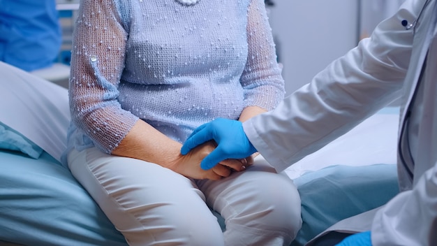 Close up van dokter handen in blauwe beschermende handschoenen comfortin een gestresste oude vrouw zittend op ziekenhuisbed in moderne privékliniek. Wereldwijde pandemie coronavirus covid-19 gezondheidszorg pandemie