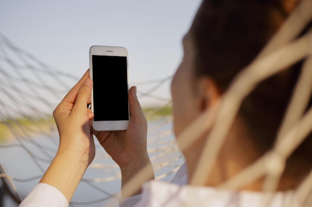 Close-up van donkerharige vrouw liggend op hangmat met behulp van sociale app op mobiele telefoon terwijl u ontspant en rust in de buurt van de rivier kopieerruimte voor reclame