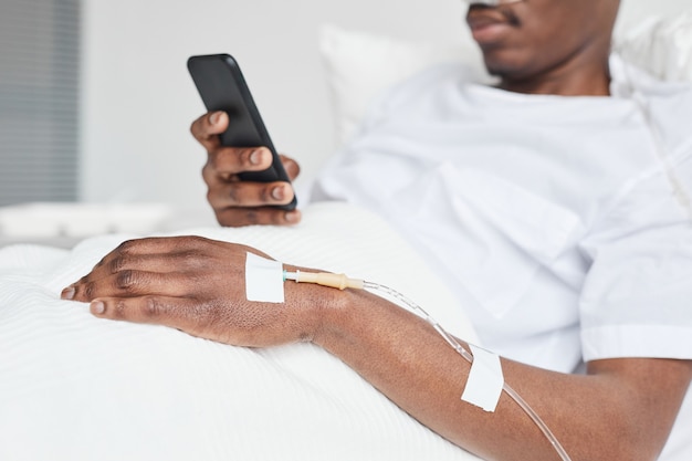 Close-up van een Afro-Amerikaanse man in ziekenhuisbed en met behulp van smartphone met focus op iv-infuuskatheter in de hand, kopieer ruimte