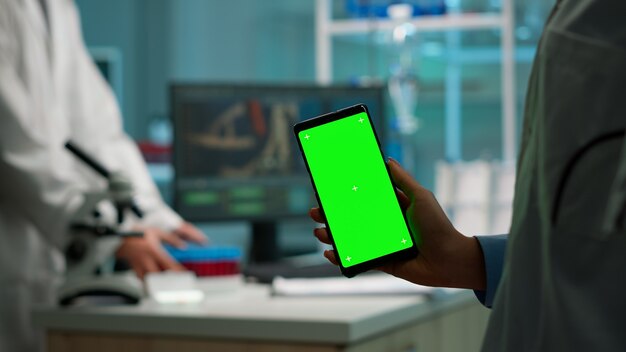 Close-up van een biochemicus die de telefoon vasthoudt en bekijkt met een chroma key-display in een biologisch laboratorium terwijl de verpleegster in een witte jas bloedmonsters brengt. Wetenschapper met behulp van telefoon met mockup, groen scherm