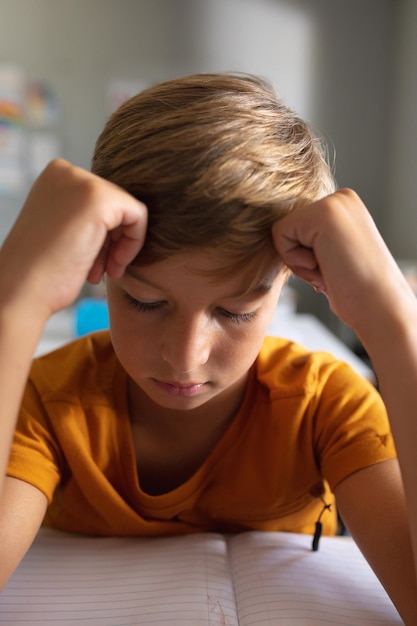 Close-up van een blanke basisschooljongen met het hoofd in de handen die aan het bureau in de klas leert. ongewijzigd, onderwijs, leren, jeugd, studeren en schoolconcept.