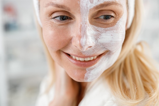 Close-up van een blanke blonde vrouw met een wit crèmemasker op de helft van haar gezicht lacht