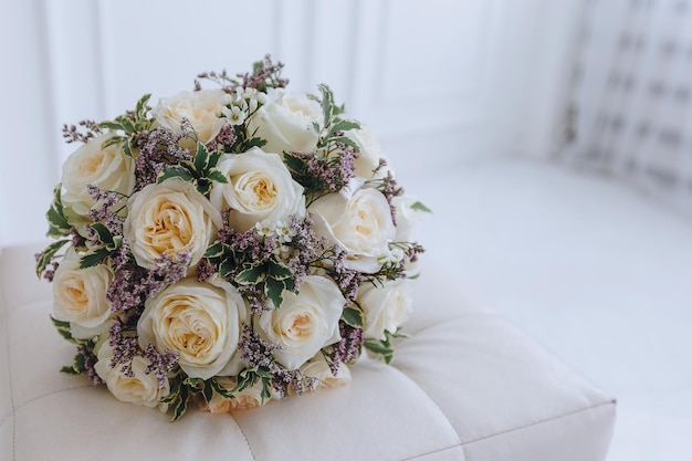 Close-up van een bruiloftsboeket van crèmekleurige rozen met paarse bloemen ligt op een witte bank in een lichte kamer