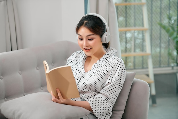 Close-up van een bruinharige casual vrouw die van muziek geniet terwijl ze een boek leest op de bank in een licht huis