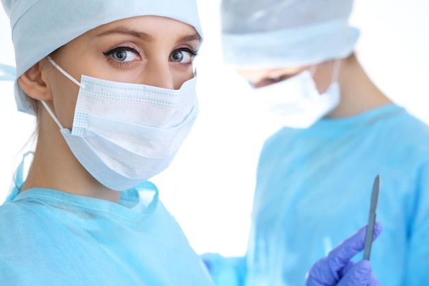 Close-up van een chirurg vrouw die naar de camera kijkt terwijl collega's op de achtergrond optreden in de operatiekamer, geïsoleerd. Chirurgie en geneeskunde concept