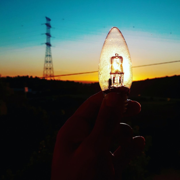 Foto close-up van een gekapte hand die een gloeilamp vasthoudt tegen de zon in de lucht tijdens de zonsondergang