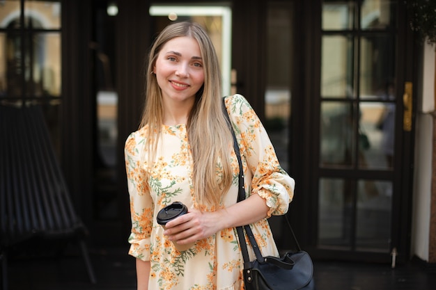 Close-up van een gelukkige blanke vrouw blonde in een jurk met een zwarte tas koffie afhaalmaaltijden drinken op een zwarte glazen achtergrond in de stad